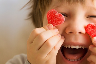 Young child holding up two red candy hearts in front of their eyes with a cheeky smile.