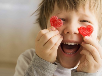 A child holding up 2 heart shaped candies next to its eyes smiling cheekishly.