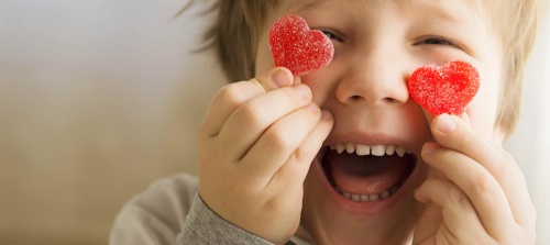 A child holding up 2 heart shaped candies next to its eyes smiling cheekishly.