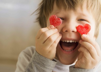 A child holding up 2 heart shaped candies next to its eyes smiling cheekishly.